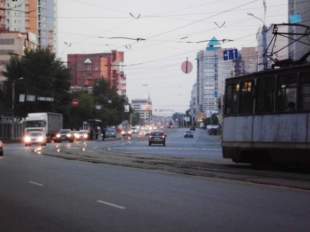 Eine belebte Stadtstraße mit Verkehr, einschließlich eines Busses und Lastwagens, Straßenlaternen, Schildern, Bäumen, Versorgungsmasten, Drähten, Gebäuden und einem klaren blauen Himmel.