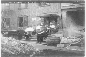 Eine Gruppe jüdischer Neujahrsverehrer steht vor einem Gebäude mit Fenstern und einer Tür, mit Stufen, die hinaufführen, in Brooklyn, New York im Jahr 1907. Links ist ein Haufen Erde zu sehen, und rechts befinden sich hölzerne Gegenstände, ein Eimer und ein Rohr. Das Bild ist in Schwarz-Weiß.