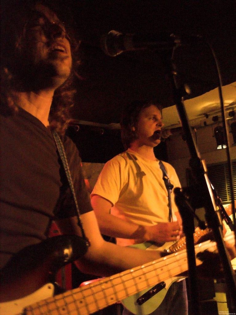 Ein Musikkonzert mit zwei Sängern: einer in einem weißen Hemd, der eine grüne Gitarre hält und ein Mikrofon in der Hand hat, und ein anderer in einem schwarzen T-Shirt, der eine Brille trägt und eine schwarze Gitarre hält, mit Scheinwerfern und anderen Instrumenten im Hintergrund.