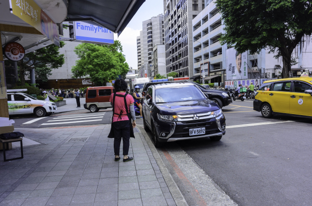 Frau geht öffentlich neben einem gelben Taxi, mit anderen Fahrzeugen auf der Straße, einem zugenagelten Gebäude und einer Bank auf der linken Seite und Bäumen und Gebäuden im Hintergrund unter einem bewölktem Himmel.