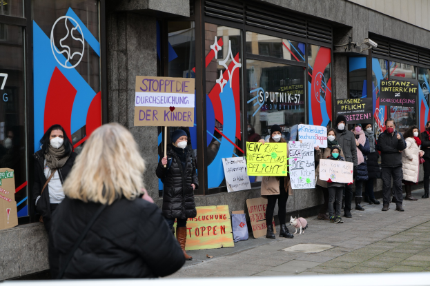 Gruppe von Menschen in Masken mit Schildern und Plakaten vor einem Gebäude mit Glaswänden und -türen, mit Kameras und einem Hund, die gegen ein deutsches Regierungsverbot von Masken in Schulen protestieren.