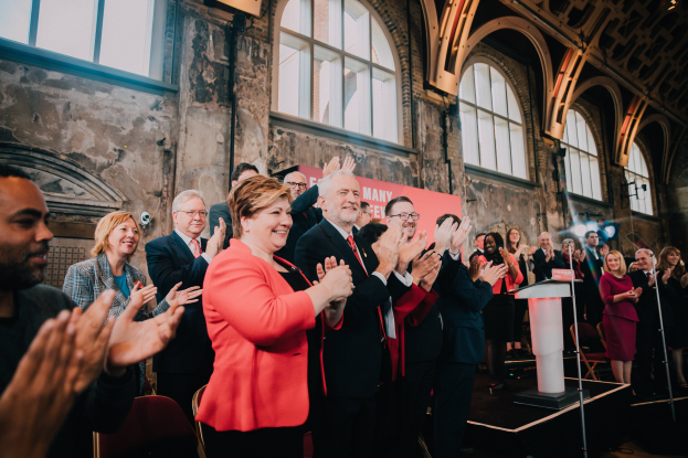 Eine Gruppe von Menschen steht vor einem Publikum und klatscht, mit einem Podium und einem Mikrofon auf der rechten Seite, Stühlen, einer Wand, einer Tafel mit der Aufschrift "Covid-19 New York City Mayor Bill de Blasio" und Fenstern im Hintergrund.