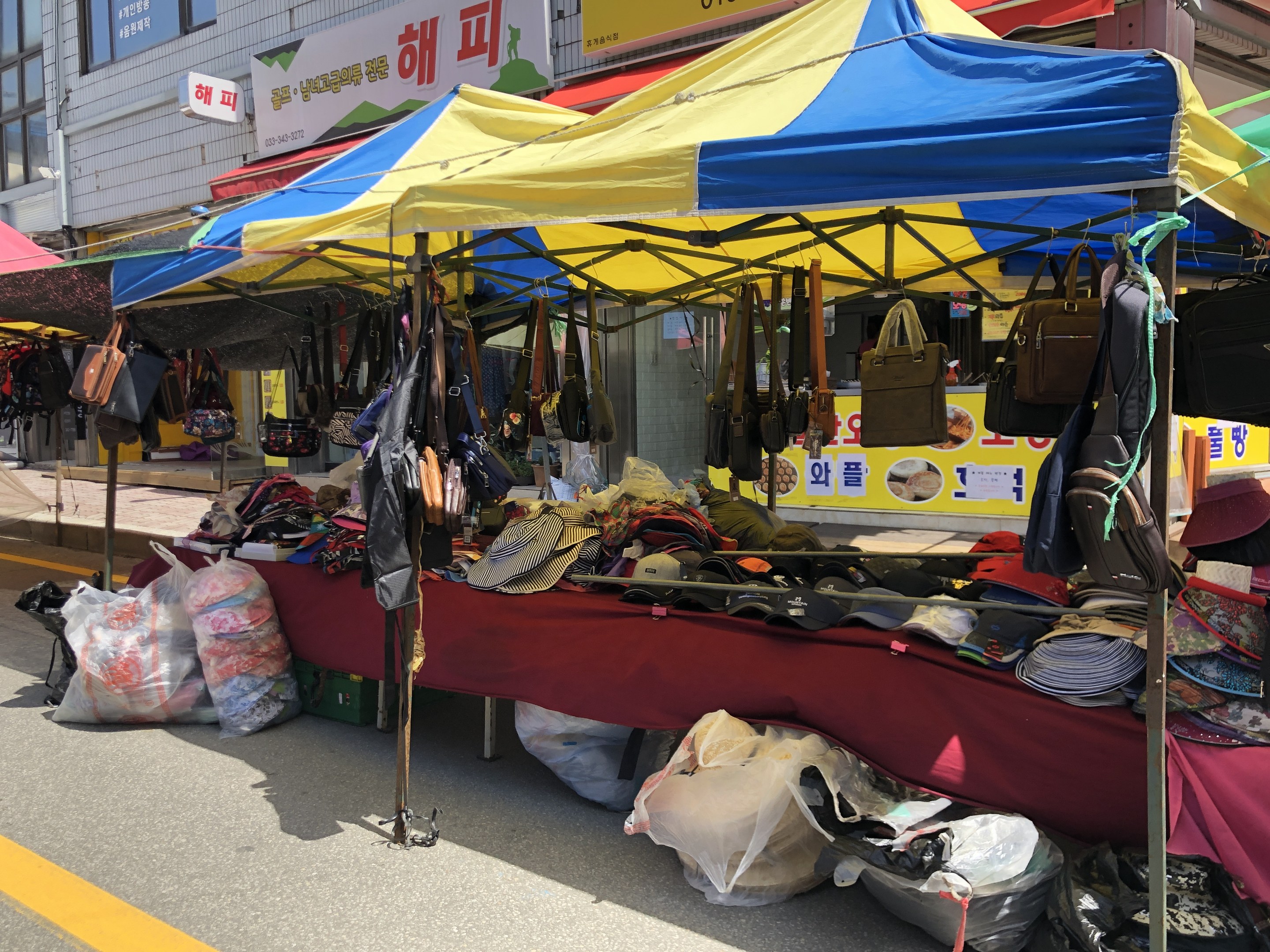 Ein Straßenmarkt mit Zelten, die Taschen, Hüte und andere Gegenstände präsentieren, neben Gebäuden mit Fenstern und Texttafeln.