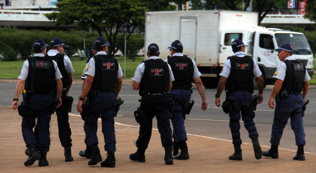 Eine Gruppe von Beamten in Uniform, einschließlich Mützen, Hemden, kugelsicheren Westen, Uhren, Hosen und Schuhen, geht auf einer Straße. Rechts daneben parkt ein Lastwagen in der Nähe eines Zauns, mit Gebäuden, Bäumen, Pflanzen, Pfählen und Bannern im Hintergrund und einer Brücke oben rechts.