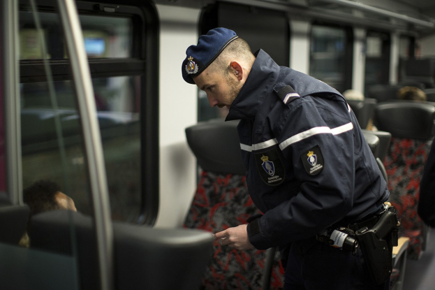 Ein Polizist in Uniform und mit Mütze steht in einem Zug, schaut auf sein Handy, mit sitzenden Passagieren und einer weiteren stehenden Person im Bild.