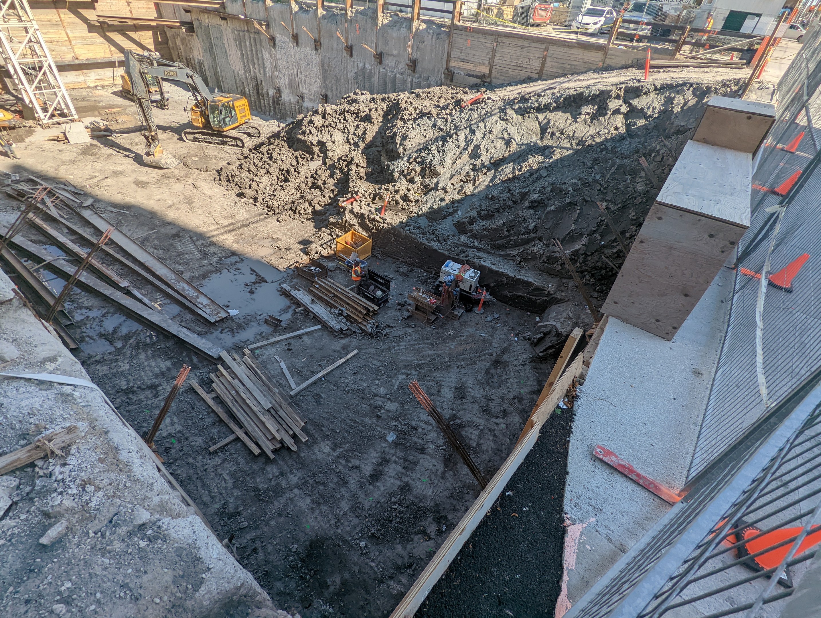 Baustelle mit einem großen Schutthaufen, Kränen, Holzstangen, einem Maschendrahtzaun auf der rechten Seite und Gebäuden, Fahrzeugen, Pfählen und Bäumen im Hintergrund unter dem Himmel.