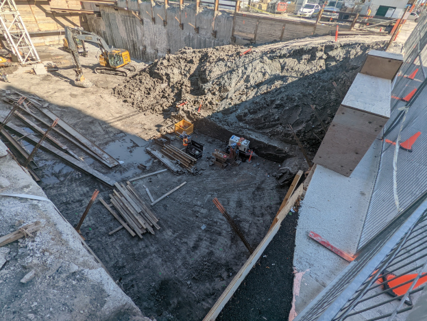 Baustelle mit einem großen Schutthaufen, Kränen, Holzstangen, einem Maschendrahtzaun auf der rechten Seite und Gebäuden, Fahrzeugen, Pfählen und Bäumen im Hintergrund unter dem Himmel.