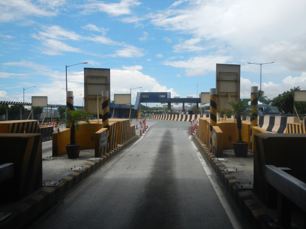 Stra├če mit einer Mautstelle, Barrieren, Verkehrskegel, Blument├Âpfe, Schilder, Stra├čenlaternen, B├Ąume und eine bew├Âlktete Himmel.