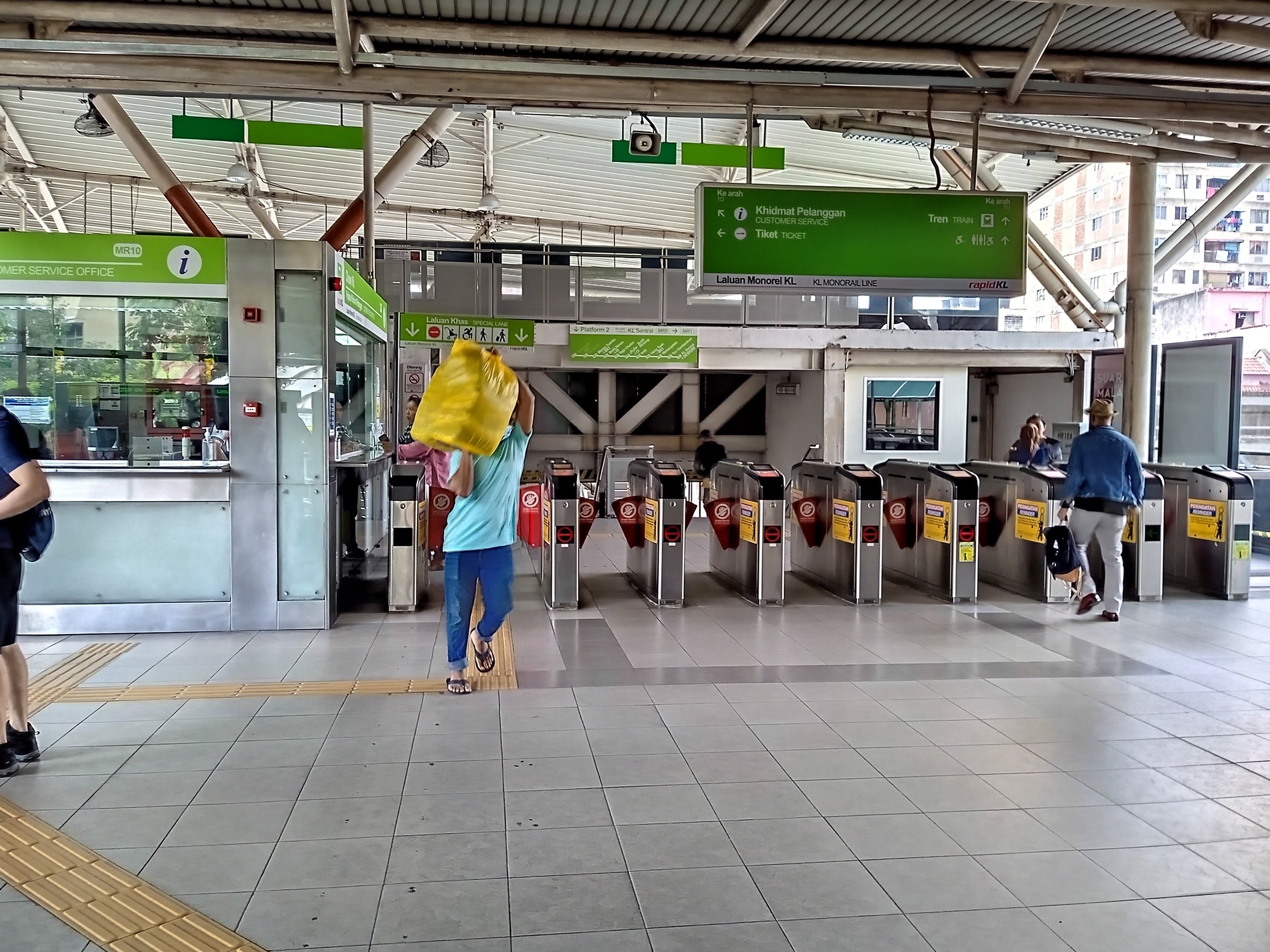 Menschen gehen durch einen Bahnhof mit Metalldetektoren, Hinweisschildern, Metallstangen, Deckenleuchten und Gebäuden mit Fenstern im Hintergrund.