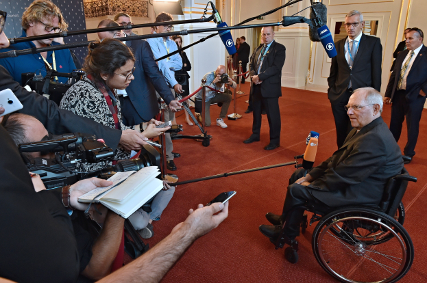 Ein Mann im Rollstuhl wird von Reportern mit Kameras, Mikrofonen und Handys umgeben, mit einem Banner, Scheinwerfern und einer Wand im Hintergrund.