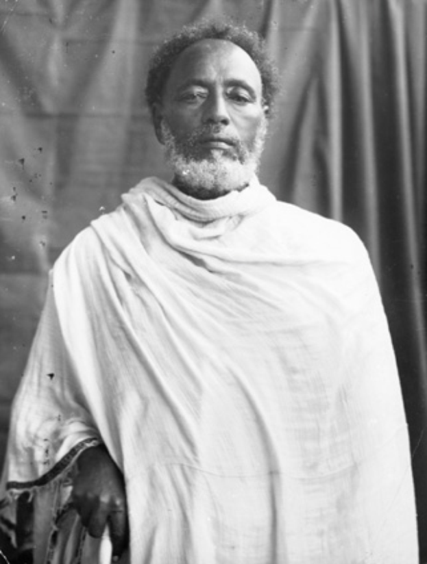 Ein Schwarz-Weiß-Foto eines bärtigen Mannes in einem Schal vor einem Vorhang, der angeblich Mahatma Gandhi ist.