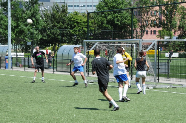 Menschen in Kleidung, die Fußball auf einem Feld mit einem Zaun und einem Fußballtor in der Mitte spielen, ein Pfosten und ein Fußball oben links und Bäume und Gebäude im Hintergrund.
