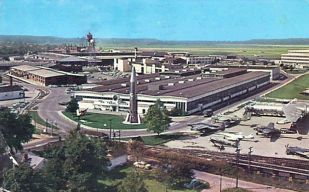 Altes Schwarz-Weiß-Foto eines Flughafens mit geparkten Flugzeugen, Gebäuden, Bäumen, Straßen, Fahrzeugen, Strommasten, Gras, Bahnschienen, Türmen, Hügeln und einem klaren blauen Himmel; Text erscheint unten.