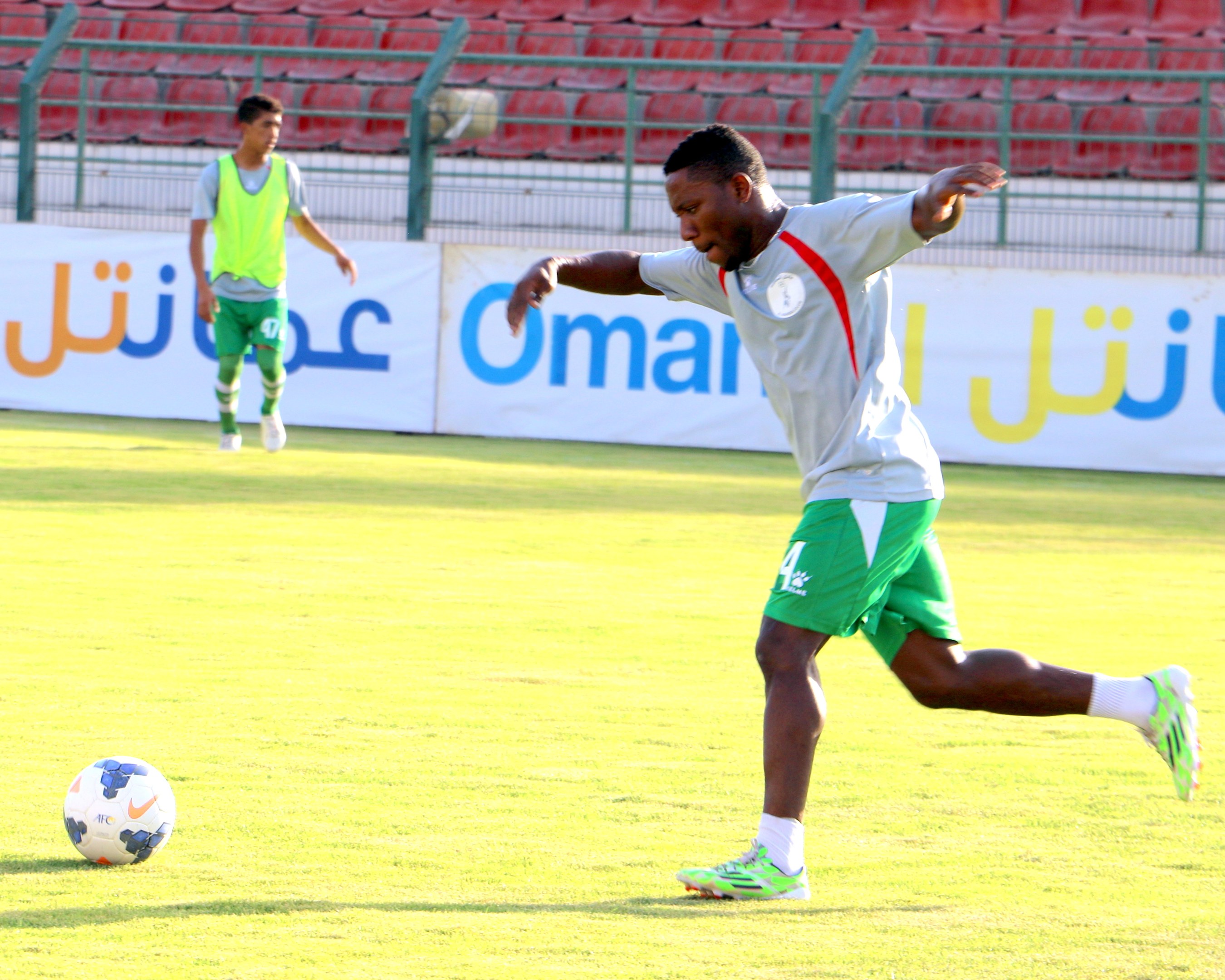 Ein Mann kickt einen Fußball auf einem Rasenfeld in einem Stadion mit zwei Personen in der Nähe und Bannern, Geländern und Stühlen im Hintergrund.