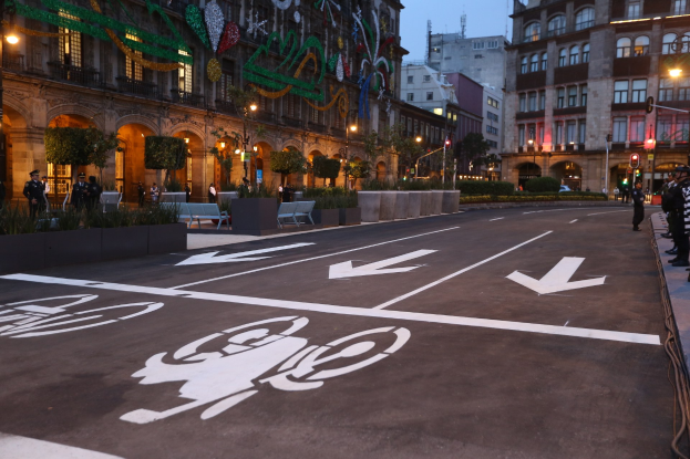 Stadtstraße mit einem Fahrradweg, umgeben von Gebäuden, Straßenmöbeln, Fußgängern und Grünflächen unter einem klaren blauen Himmel.