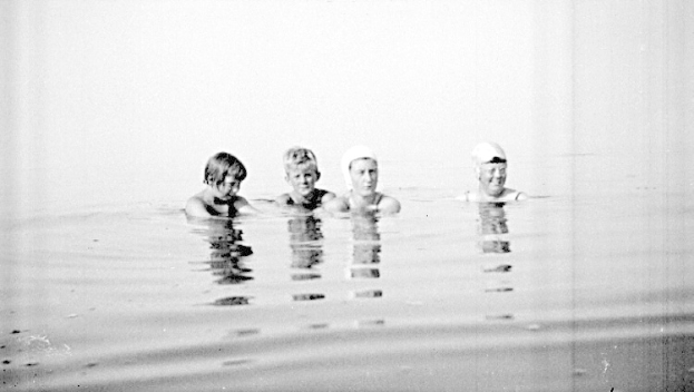 Schwarzes und weißes Foto von vier Kindern mit Schwimmkappen, die gemeinsam in einem Pool schwimmen.