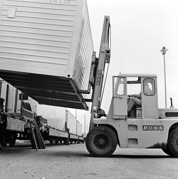 Ein Mann bedient einen Gabelstapler, der einen Container trägt und eine Straße mit anderen Fahrzeugen befährt, unter einem klaren Himmel mit Masten im Hintergrund.