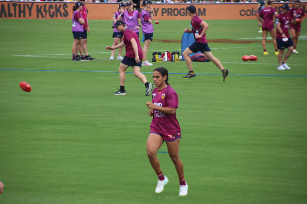 Gruppe von Frauen, die Australian Rules Football auf einem grünen Feld spielen, mit verstreuten Bällen und einem Banner im Hintergrund, einige tragen Kappen und Turnschuhe.