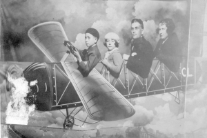 Vier Personen in einem Flugzeug sitzend, eine hält eine Waffe, in Schwarzweiß.