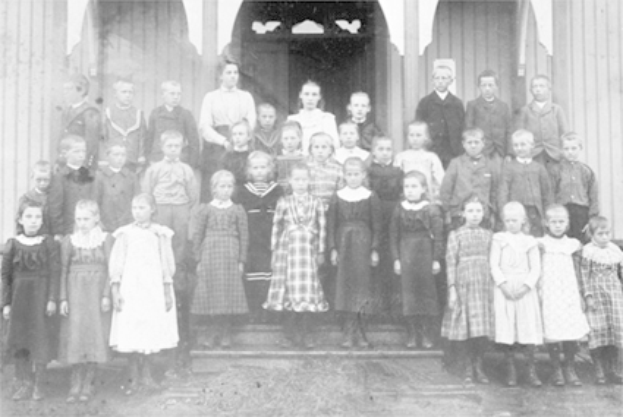 Ein Schwarz-Weiß-Foto einer Gruppe von Kindern, die vor einem Gebäude mit einer Tür und Säulen stehen, mit der Aufschrift "Erste Klasse" unten.