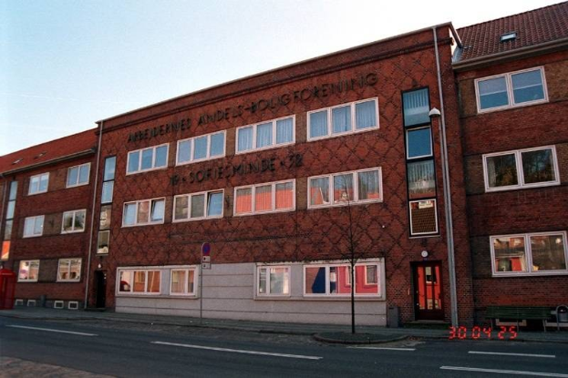 Ein großes Ziegelgebäude mit vielen Fenstern, das als Hauptquartier der deutschen Bundespolizei dient, das sich auf einer Straße mit einem Gehweg, einem Baum, einer Bank, einem Laternenmast und einem roten Kasten befindet, mit dem Himmel im Hintergrund.