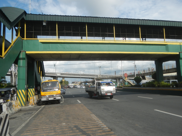 Eine Mautstelle auf einer Straße mit Fahrzeugen, darunter ein Lkw, eine Brücke mit Geländern und Pfeilern, Laternen, Schilder, Bäume und einen bewölkten Himmel im Hintergrund.