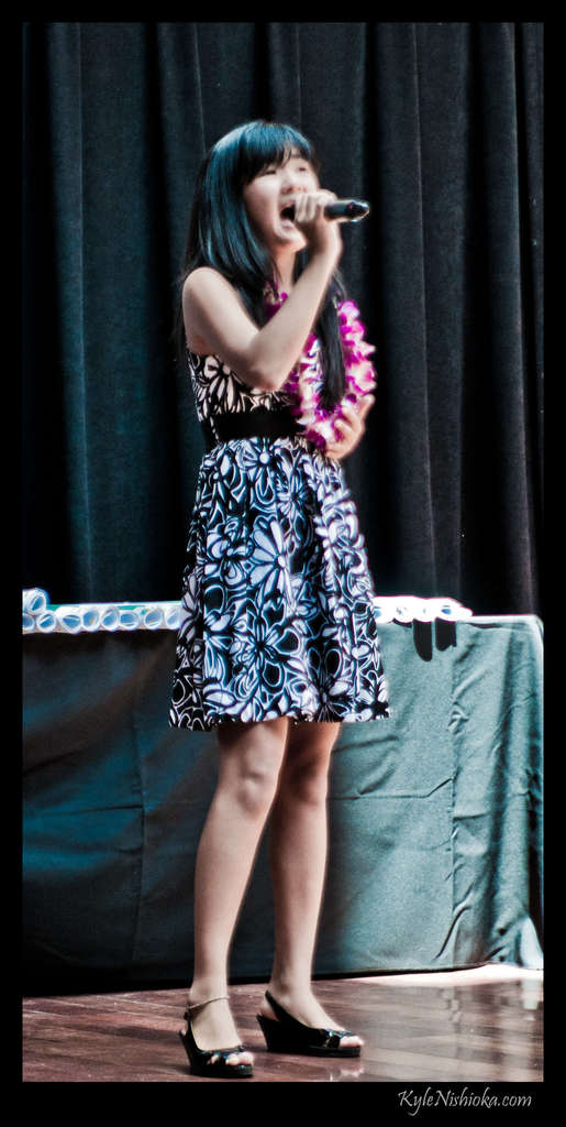 Eine Frau in einem bunten Kleid singt in ein Mikrofon auf der B├╝hne, mit einem Tisch und Papieren im Hintergrund hinter einem Vorhang.