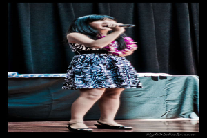 Eine Frau in einem bunten Kleid singt in ein Mikrofon auf der B├╝hne, mit einem Tisch und Papieren im Hintergrund hinter einem Vorhang.