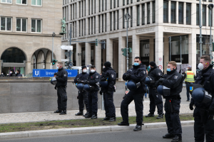 Eine Gruppe von Polizisten in schwarzen Uniformen und Masken steht vor einem Berliner Gebäude mit Glasfenstern und Säulen, einige halten Helme, während Laternenpfähle, Verkehrsampeln und andere Menschen im Hintergrund zu sehen sind.