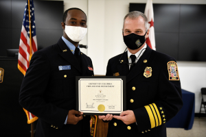 Zwei Männer in Feuerwehranzügen stehen nebeneinander, tragen Masken und halten ein Zertifikat, mit Fahnen und Möbeln im Hintergrund; der Mann links ist der Feuerwehrchef des Bezirks Washington und erhält eine Auszeichnung.