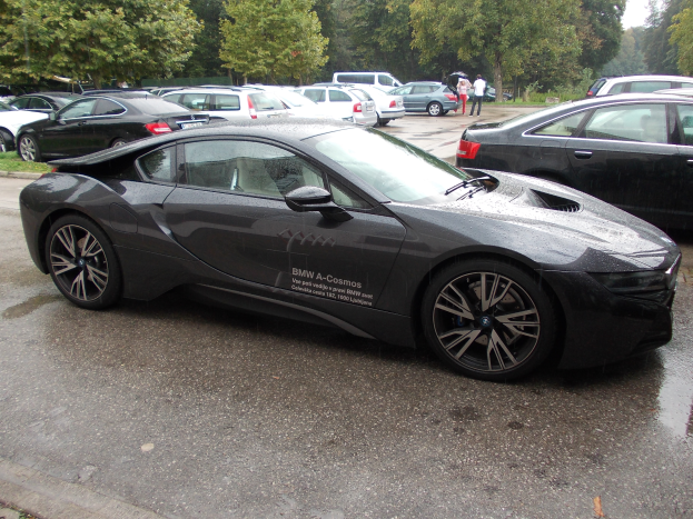 Ein schwarzer BMW i8 auf einem Parkplatz mit anderen Autos und Menschen in der Nähe, mit Bäumen und einem klaren blauen Himmel im Hintergrund.