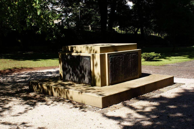 Ein Weltkriegsdenkmal steht in einem Park, umgeben von grünem Gras, Pflanzen und Bäumen mit dem Himmel darüber.