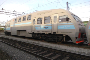 Ein diesel-elektrischer Lokomotive fährt auf Schienen neben einem anderen Zug, mit Strommasten und Kabeln im Hintergrund unter einem klaren blauen Himmel.