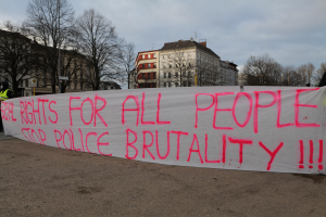 Eine Gruppe von Menschen, die auf dem Boden steht und ein Banner hält, auf dem 'Rechte für alle Menschen Stoppt Polizeigewalt' steht, mit einem Straßenschild, einem Schild, Bäumen, Gebäuden mit Fenstern und einem bewölkten Himmel im Hintergrund.