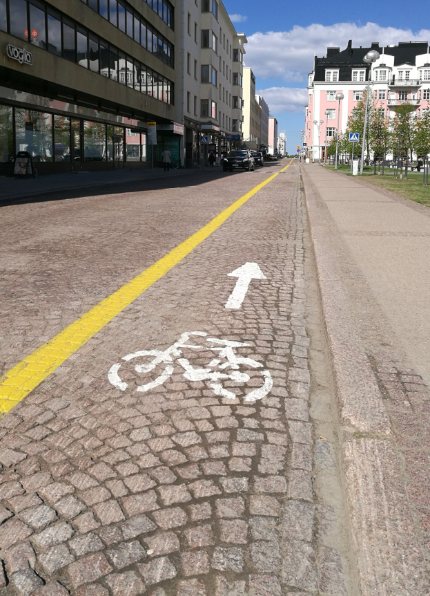 Stadtstraße mit markierter Fahrradspur, Fahrzeuge, Gebäude, Laternen, Bäume und bewölkter Himmel.