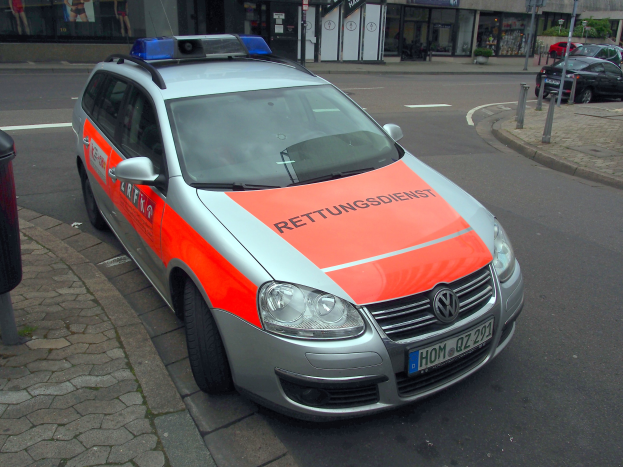 Polizeiauto am Straßenrand neben einem Müllcontainer auf dem Gehweg geparkt, mit Gebäuden und Bäumen im Hintergrund.
