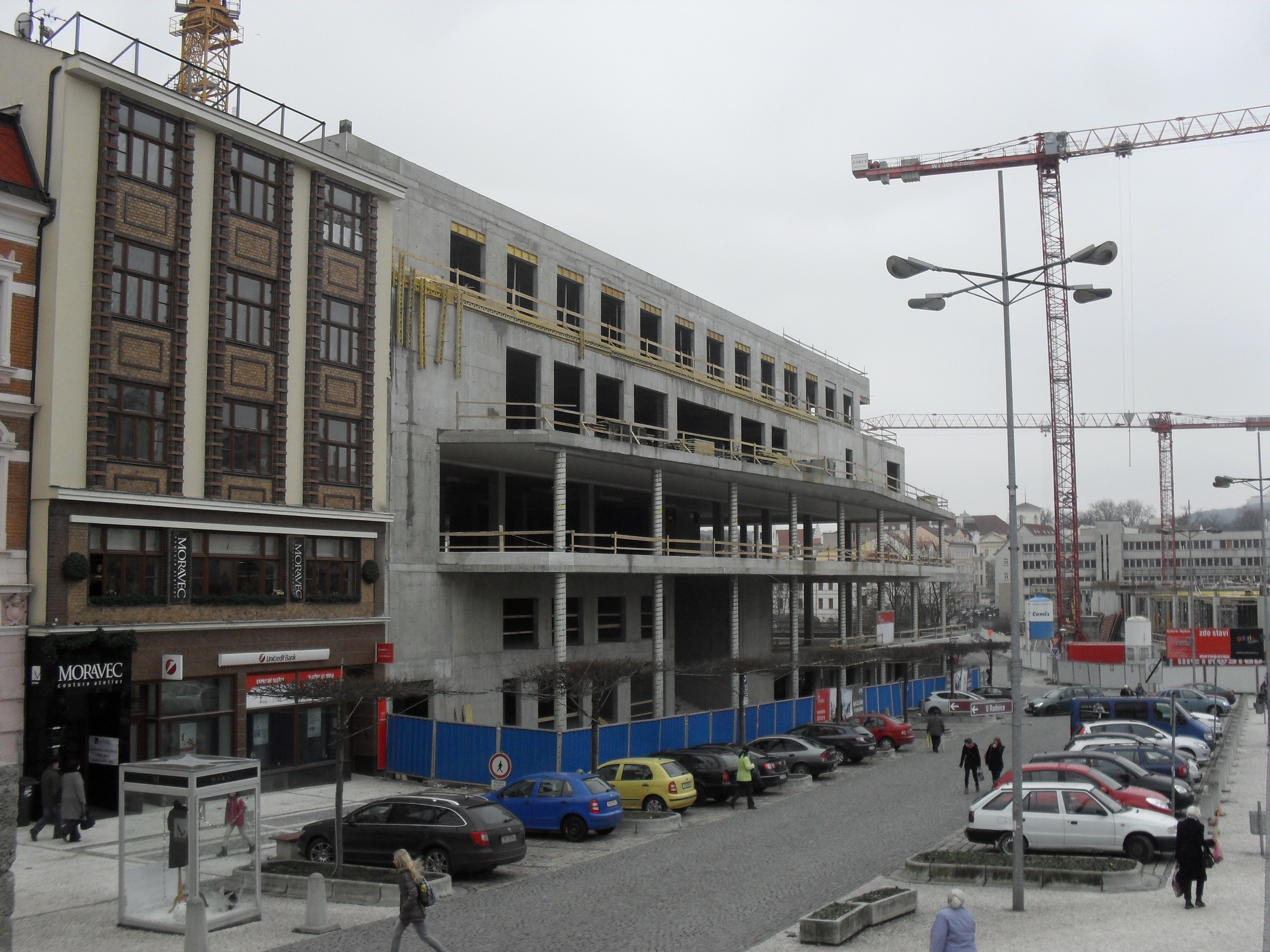 Großbau im Bau in städtischer Umgebung mit Straßeninfrastruktur, Fahrzeugen, Fußgängern, Bäumen und wolkenlosem Himmel.