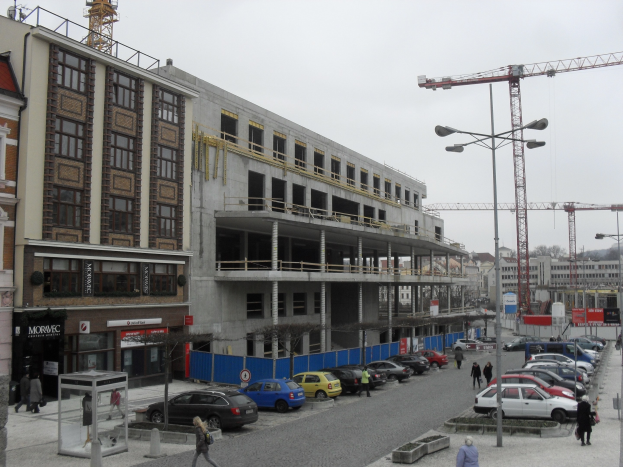 Großbau im Bau in städtischer Umgebung mit Straßeninfrastruktur, Fahrzeugen, Fußgängern, Bäumen und wolkenlosem Himmel.