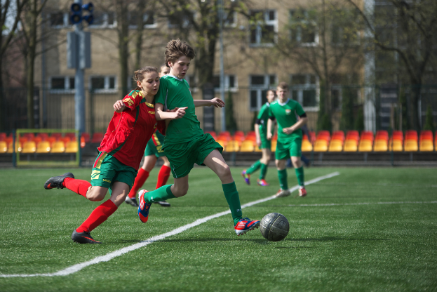 Eine Gruppe von Spielern spielt Fußball auf einem grünen Feld, das von Bäumen umgeben ist, mit ein paar Gebäuden und Stühlen für die Zuschauer im Hintergrund.
