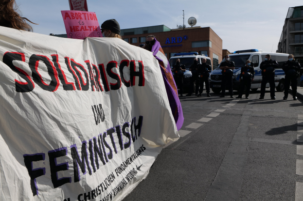 Eine Gruppe von Menschen marschiert auf einer Straße, hält ein Banner mit der Aufschrift "Solidarität und Feminismus", mit parkenden Fahrzeugen, Gebäuden, einer Schüsselantenne und einem klaren blauen Himmel im Hintergrund.