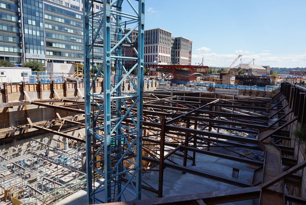Das Bild zeigt eine Baustelle mit Gebäuden und Büros links, Eisenstangen und Maschinen rechts und verschiedene Geräte und Kräne in der Mitte, unter einem Himmel in der oberen rechten Ecke.