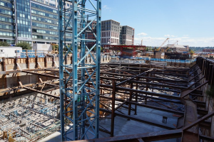 Das Bild zeigt eine Baustelle mit Gebäuden und Büros links, Eisenstangen und Maschinen rechts und verschiedene Geräte und Kräne in der Mitte, unter einem Himmel in der oberen rechten Ecke.
