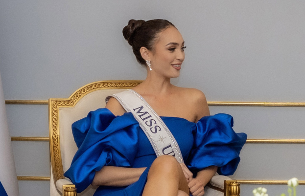 Frau in einem blauen Kleid und einer "Miss Universe 2019"-Schärpe, die auf einem Stuhl sitzt, lächelt und von Blumen umgeben ist, mit einer Wand im Hintergrund.