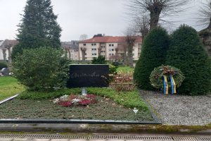 Ein Weltkriegsdenkmal für Opfer steht in einem Park, geschmückt mit einem Kranz und umgeben von Grün, mit Gebäuden im Hintergrund.