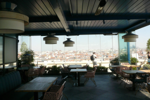 Restaurant-Innenraum mit Tischen, Stühlen und Deckenleuchten, mit Blick auf eine Stadtlandschaft aus Gebäuden, Bäumen und einem klaren blauen Himmel, mit einigen Gästen an den Tischen und Stehenden.