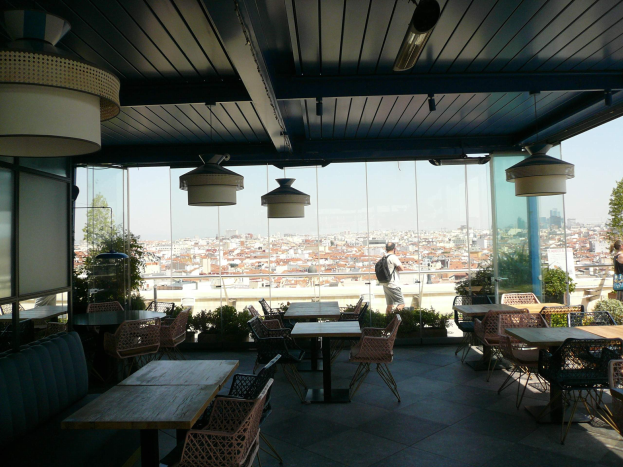 Restaurant-Innenraum mit Tischen, Stühlen und Deckenleuchten, mit Blick auf eine Stadtlandschaft aus Gebäuden, Bäumen und einem klaren blauen Himmel, mit einigen Gästen an den Tischen und Stehenden.