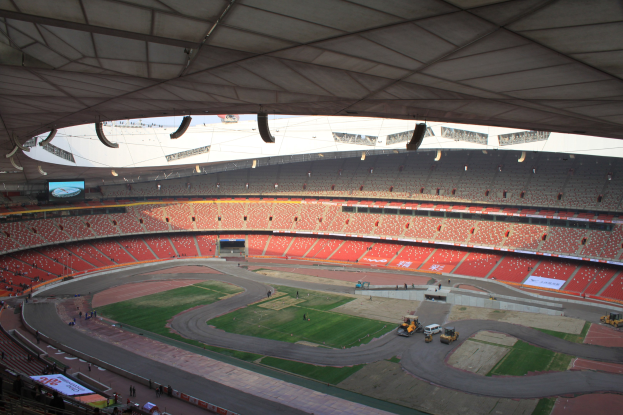 Großes Stadion voller Zuschauer, Fahrzeuge auf der Straße, grüner Boden, Texttafeln, Geländer, Deckenbeleuchtung und ein Bildschirm.