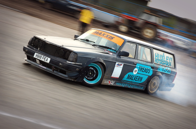 Ein Rennwagen mit Qualm, der mit hoher Geschwindigkeit auf einer Rennstrecke fährt, umgeben von anderen Fahrzeugen und Zuschauern.