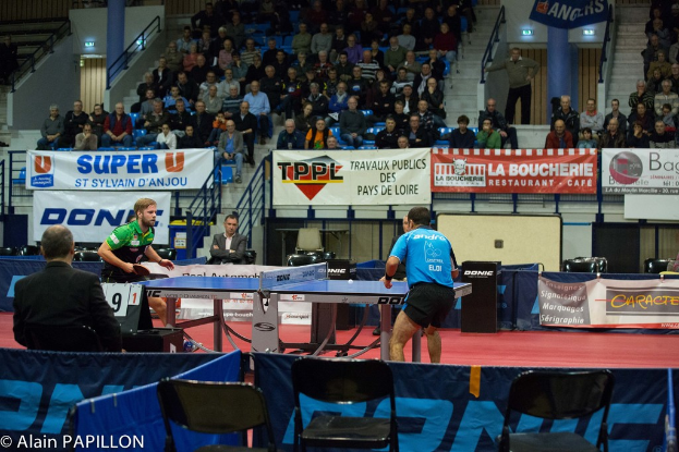 Zwei Männer spielen Tischtennis drinnen, mit mehreren Zuschauern, die stehen und sitzen, einigen leeren Stühlen und Werbeplakaten, mit Text in der unteren linken Ecke.