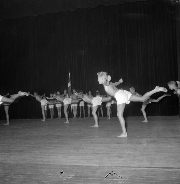 Schwarzes Foto einer Gruppe, die auf einer Bühne Gymnastik macht, mit einem Vorhang und einer Flagge im Hintergrund.
